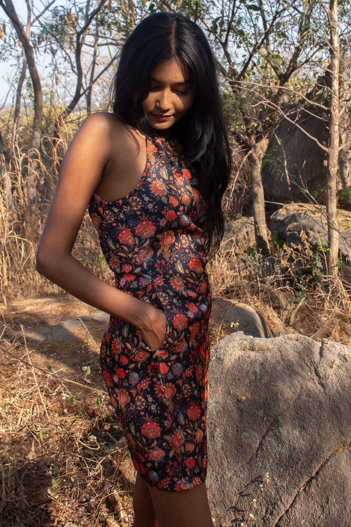 Woman in a floral dress standing in a natural setting with trees and rocks.