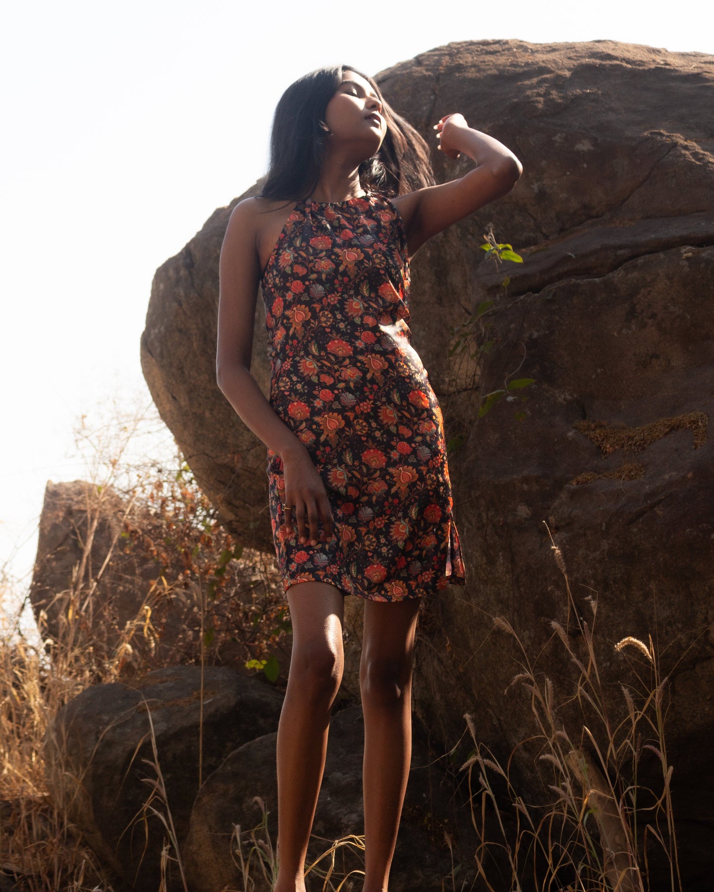 Woman in a floral dress standing on a rock with large rocks and dry grass in the background