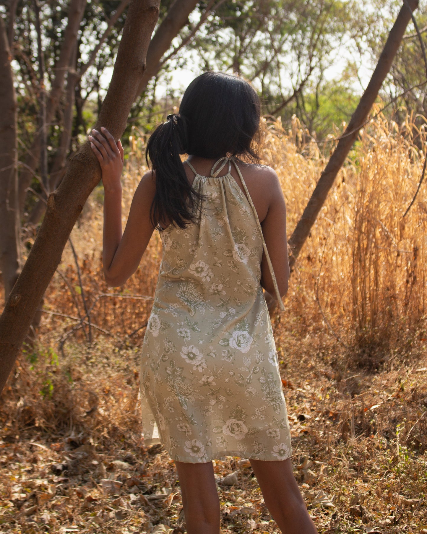 Woman in a floral dress standing in a natural setting with trees and dry grass.