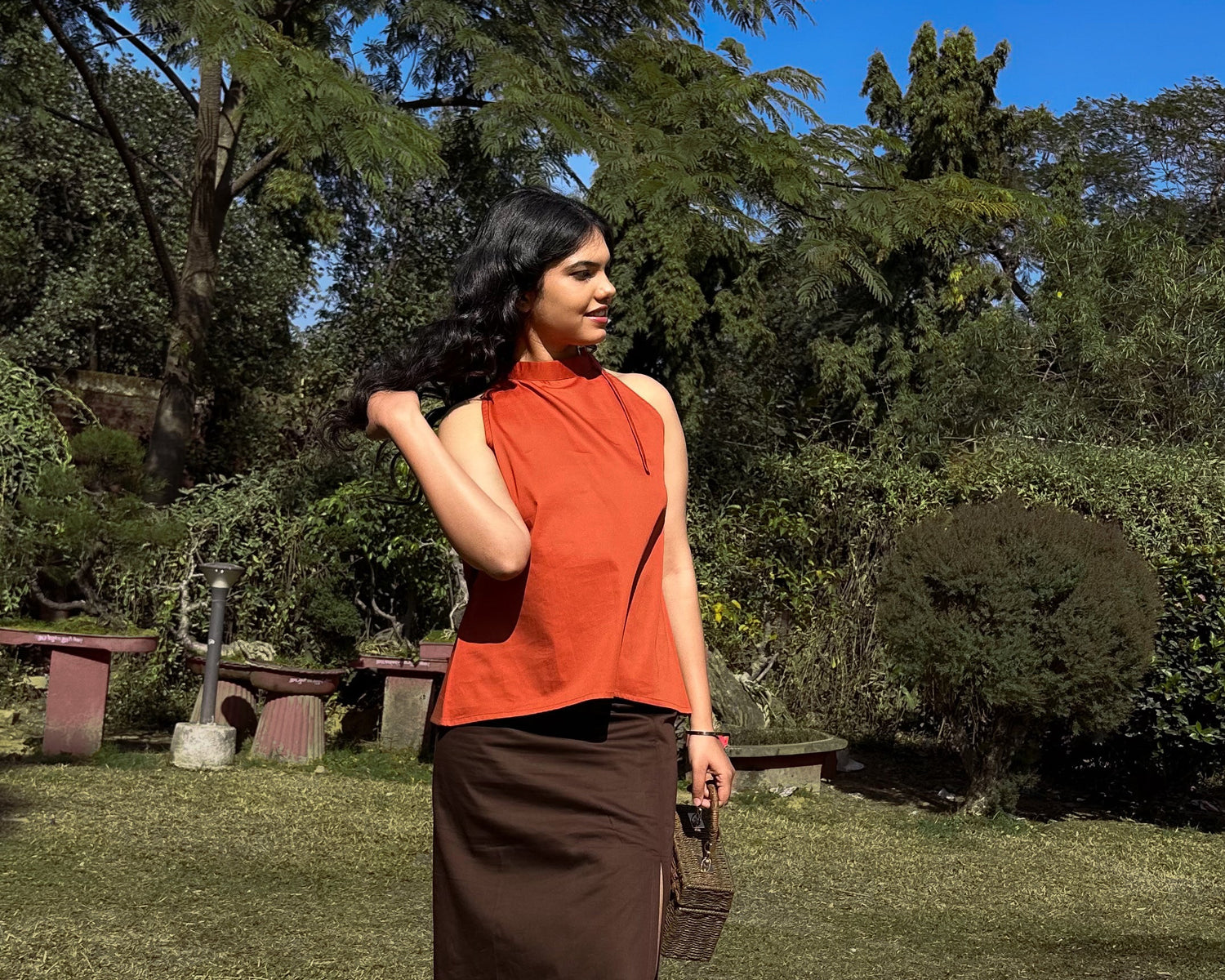 Woman in an orange top and brown skirt standing in a park with trees and grass.
