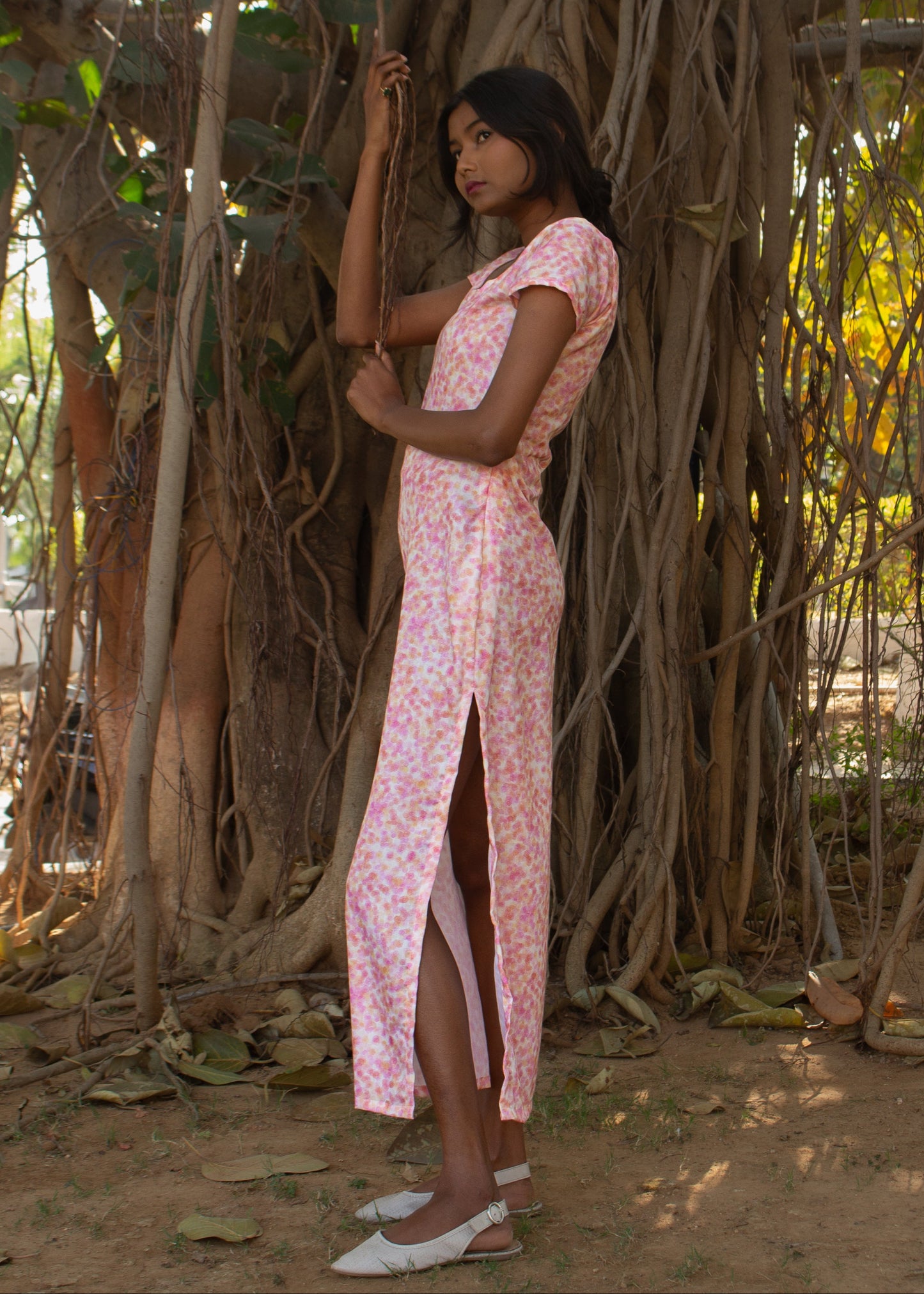 Woman in a pink dress standing under a large tree with intricate root system.