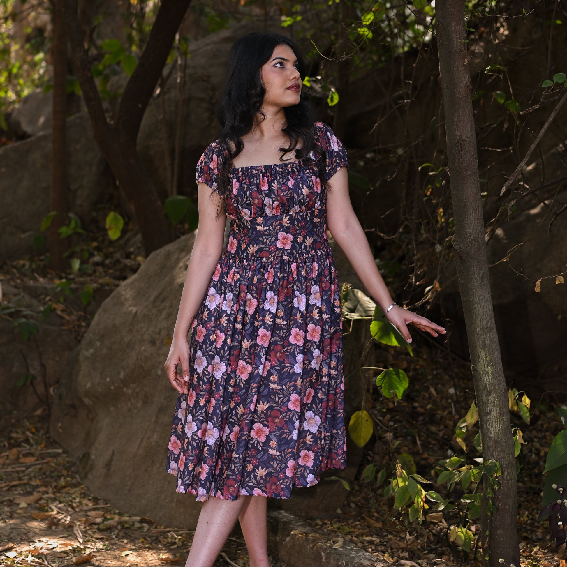 Woman in a floral dress standing in a natural setting with trees and rocks.