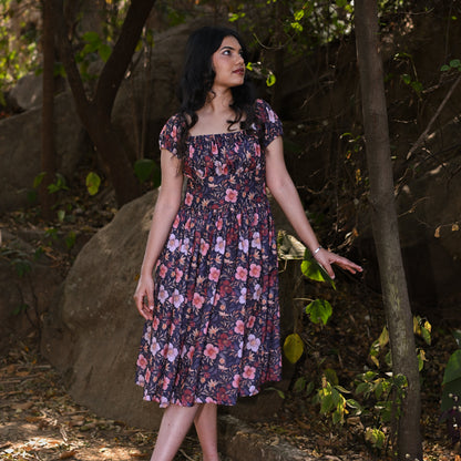 Woman in a floral dress standing in a natural setting with trees and rocks.