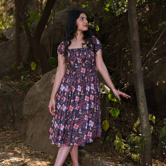 Woman in a floral dress standing in a natural setting with trees and rocks.