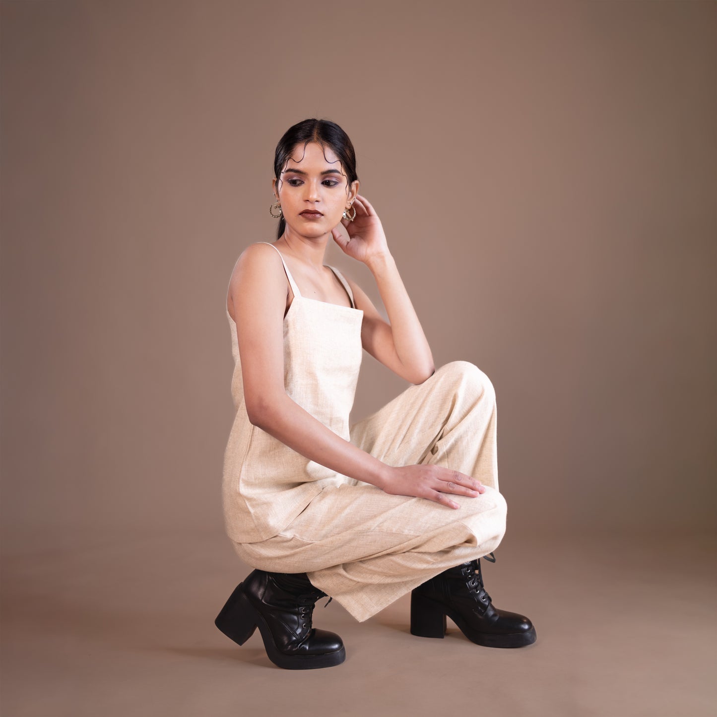 Woman in a beige top and trouser with black boots posing against a brown background