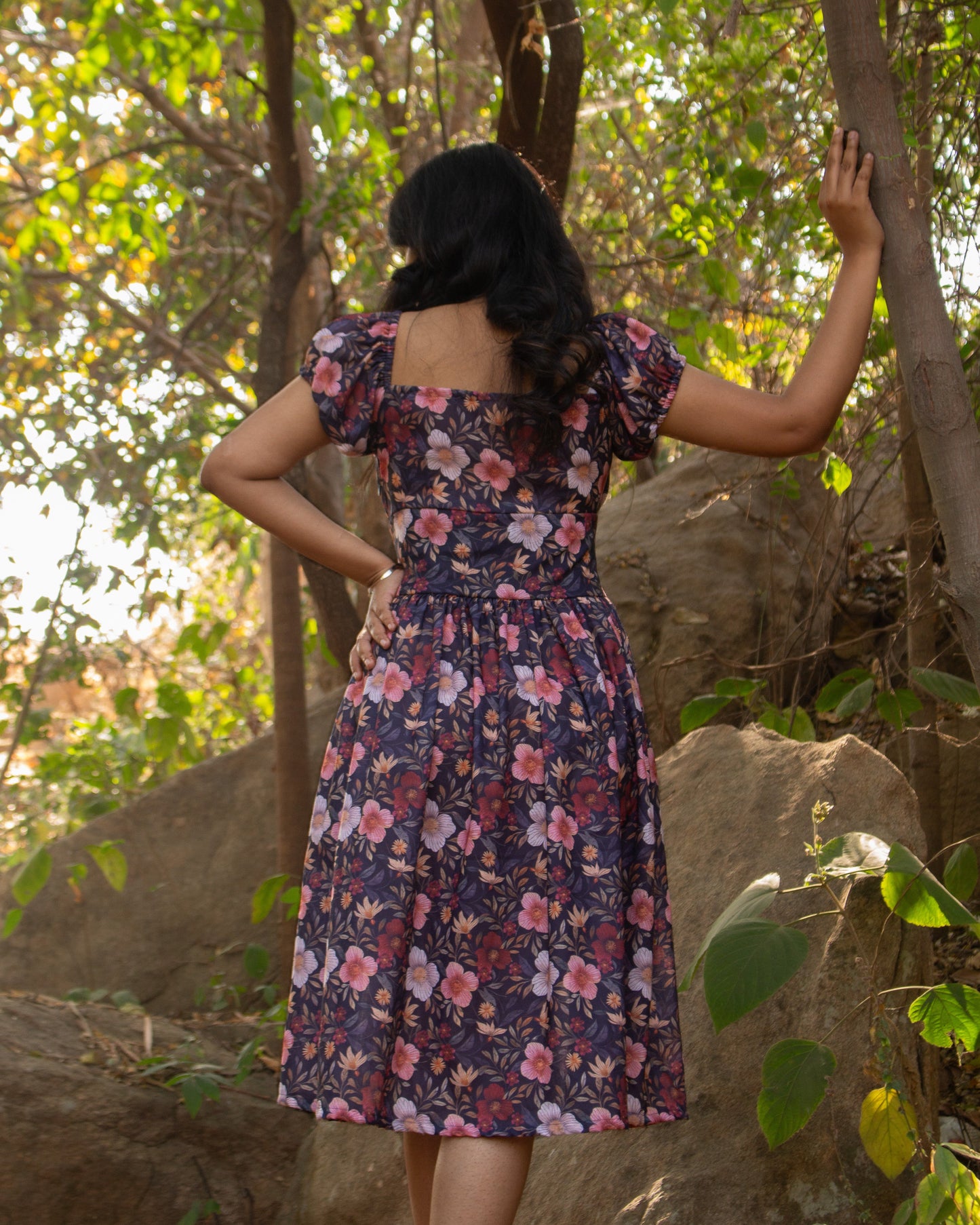 Woman in a floral dress standing in a forested area with trees and rocks.