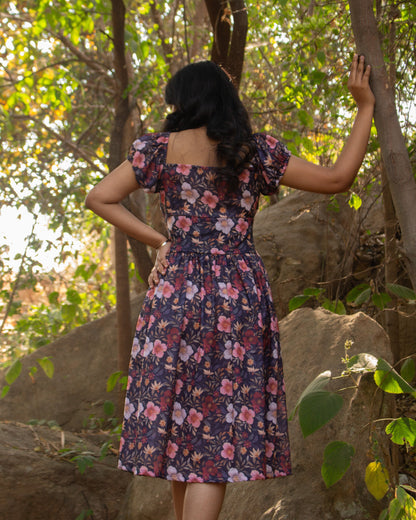 Woman in a floral dress standing in a forested area with trees and rocks.