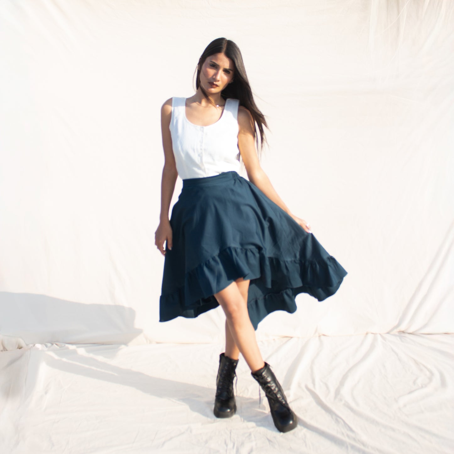 Woman wearing a white top and teal skirt with black boots on a light background