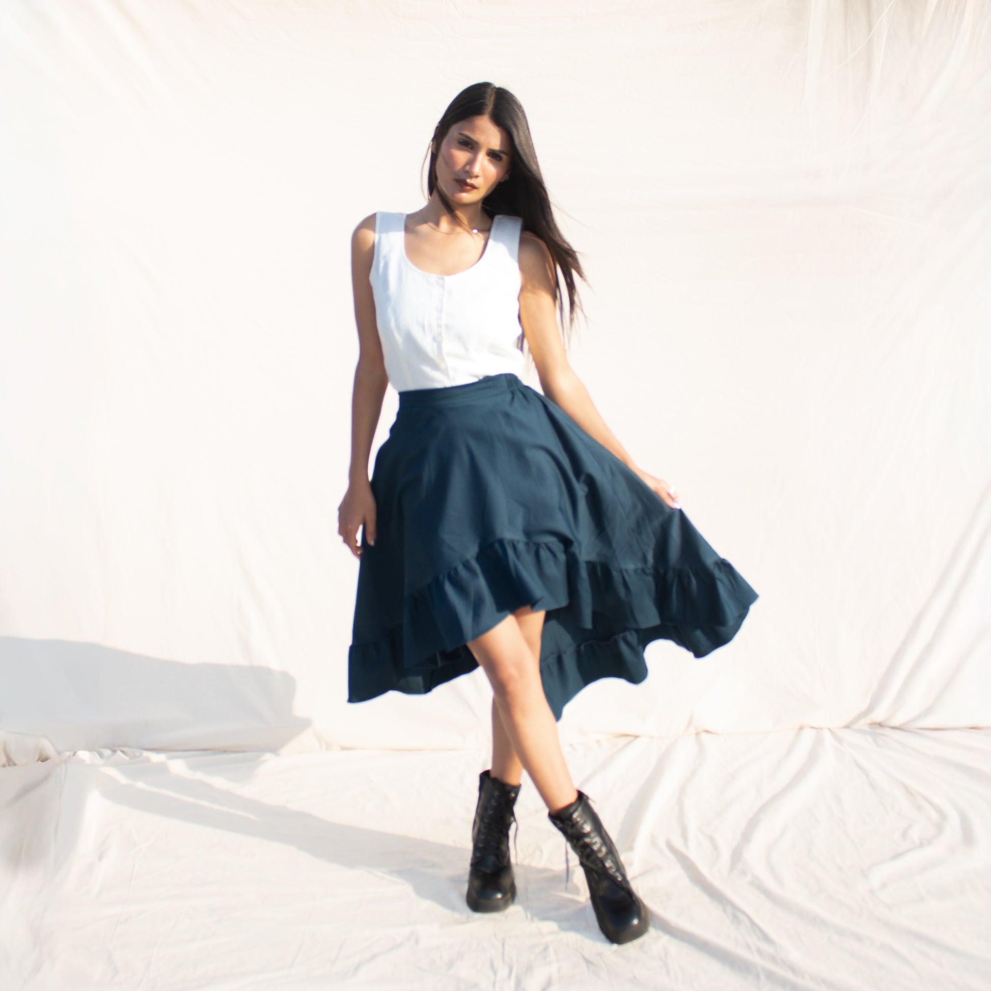 Woman wearing a white top and teal skirt with black boots on a light background