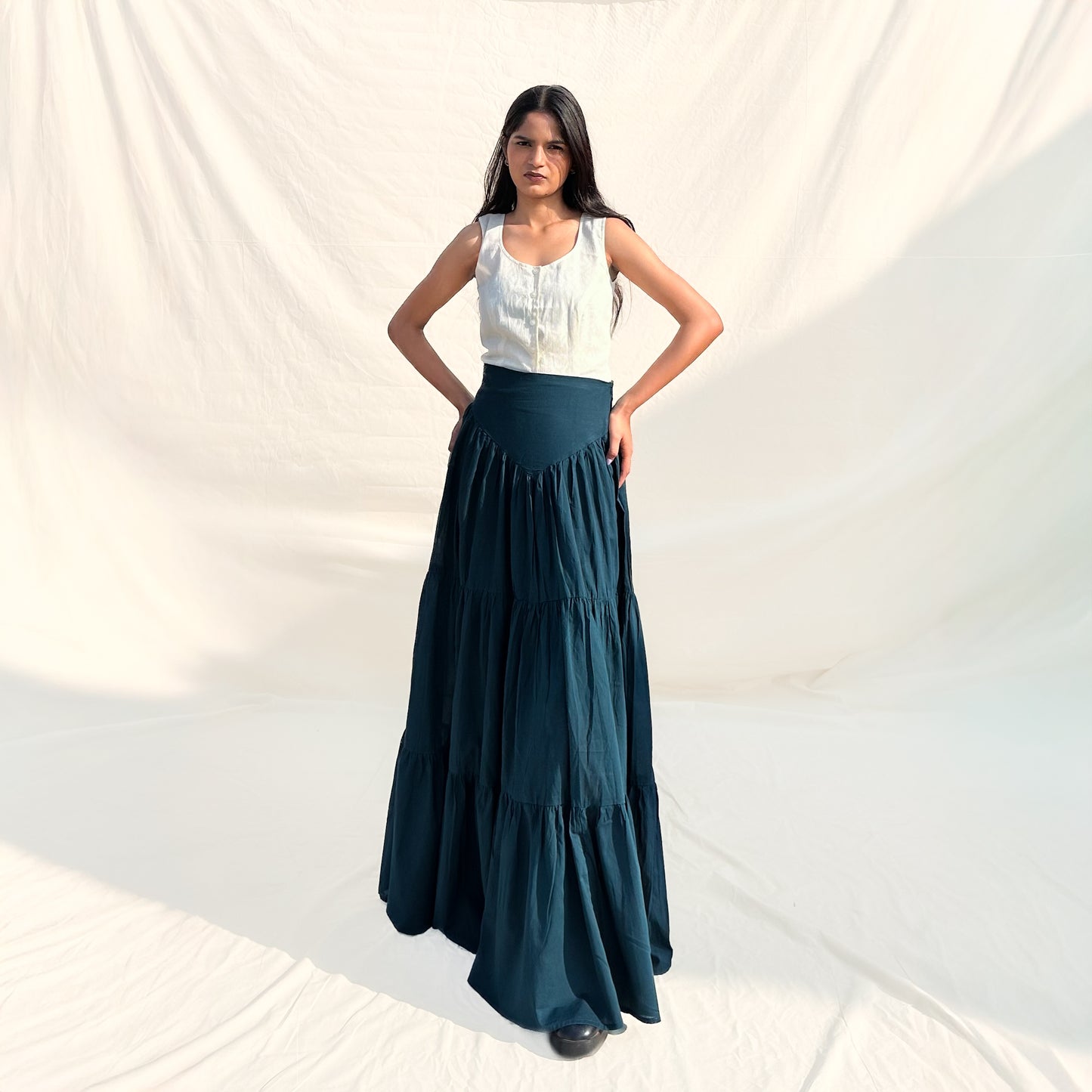 Woman wearing a white top and dark green tiered skirt against a plain background