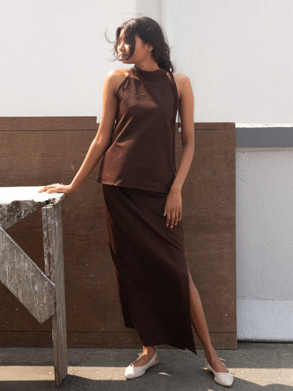 Woman wearing a brown sleeveless top and long skirt standing against a white wall.