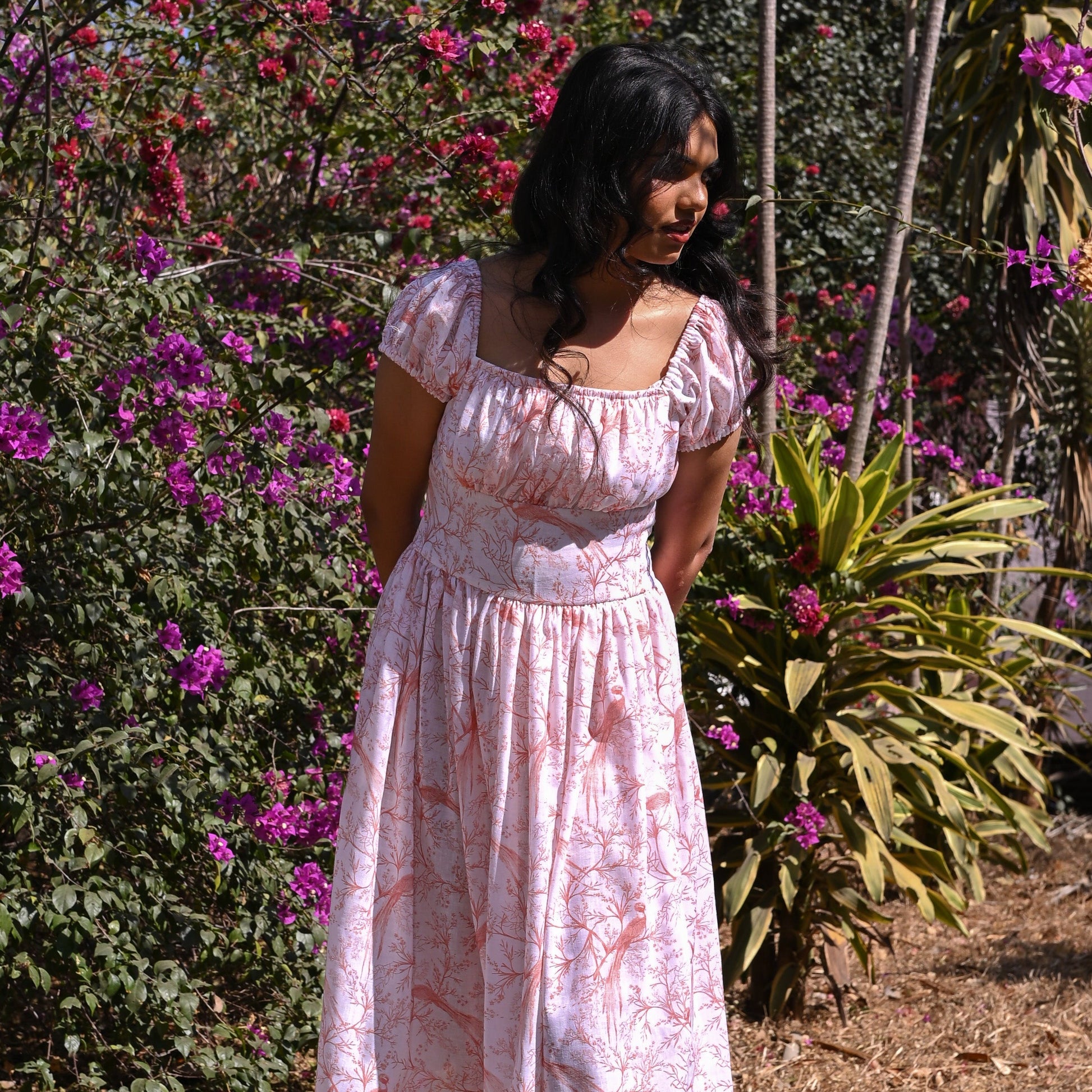 Woman in a pink floral dress standing in a garden with pink flowers and green plants.